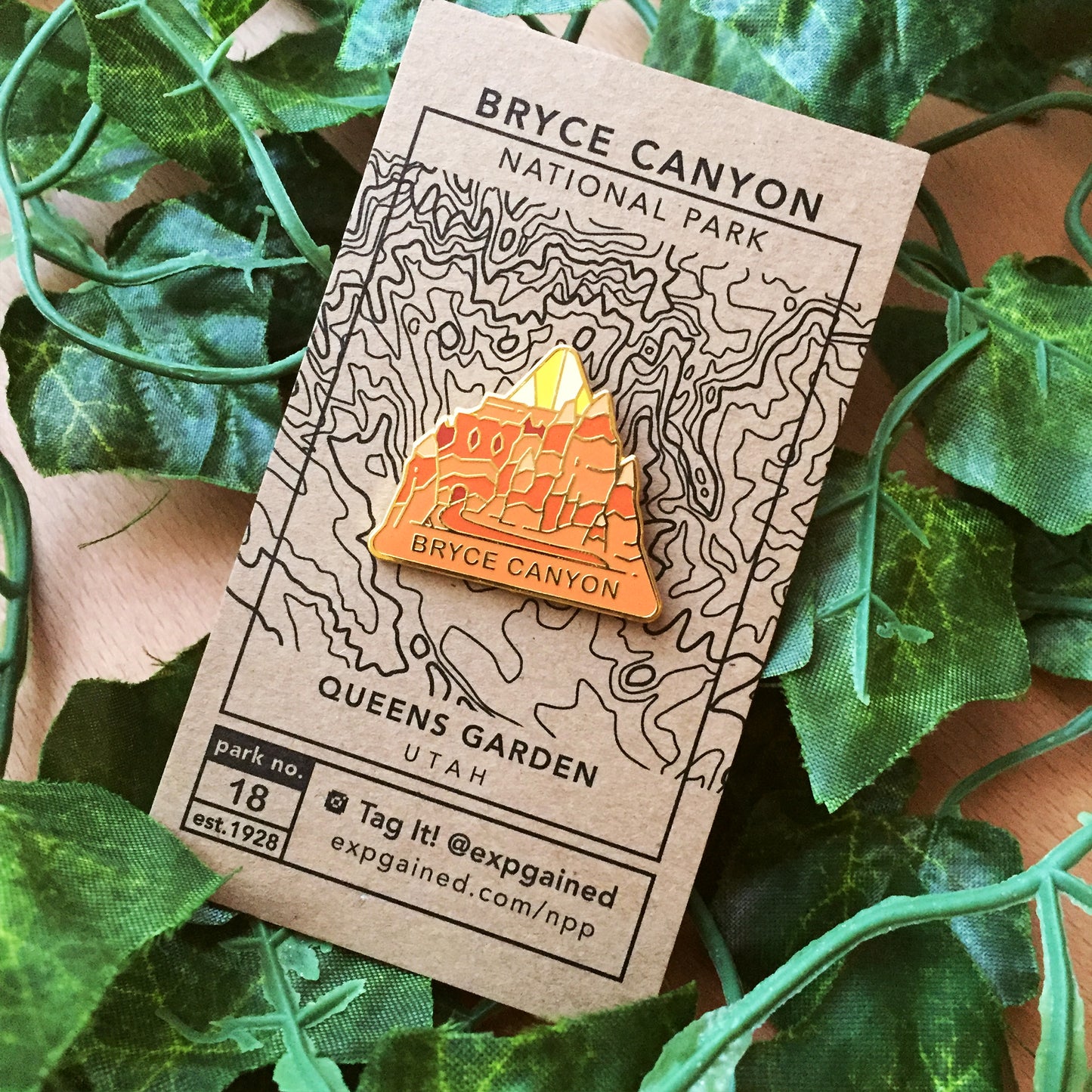 Triangle Bryce Canyon national park enamel pin featuring a view from the beehive hike on a brown business card size backing card with a topo map of the Queens Garden.