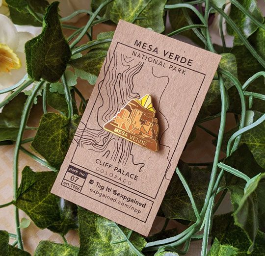 Triangle Mesa Verde national park enamel pin featuring a view from the beehive hike on a brown business card size backing card with a topo map of cliff palace.