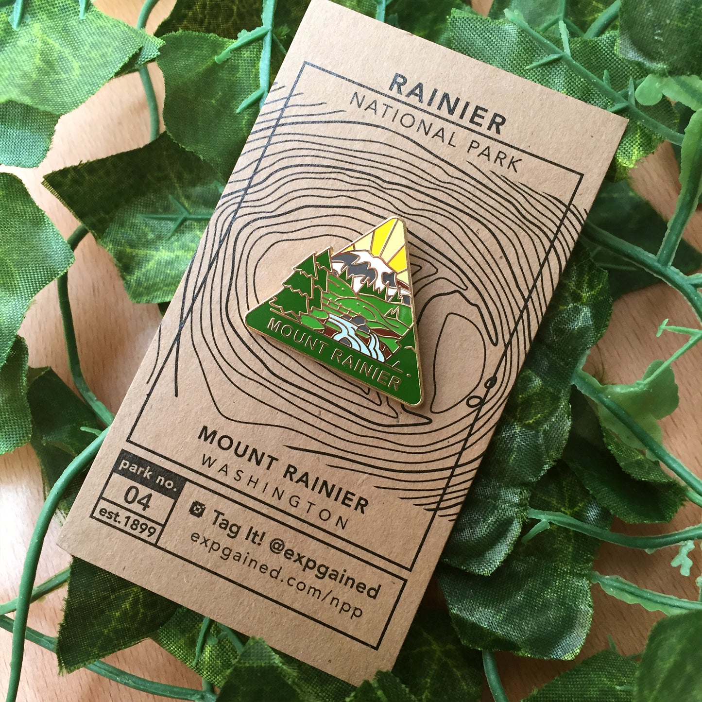 Triangle Mount Rainier national park enamel pin featuring a view from the beehive hike on a brown business card size backing card with a topo map of Mount Rainier.